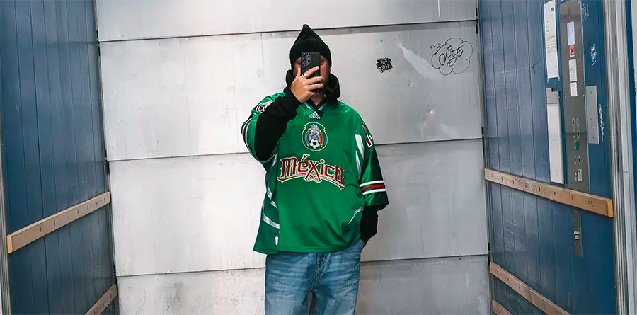 A person in a green Mexico jersey and jeans takes a selfie in an industrial elevator. The mood is casual, and the background is metallic with graffiti.