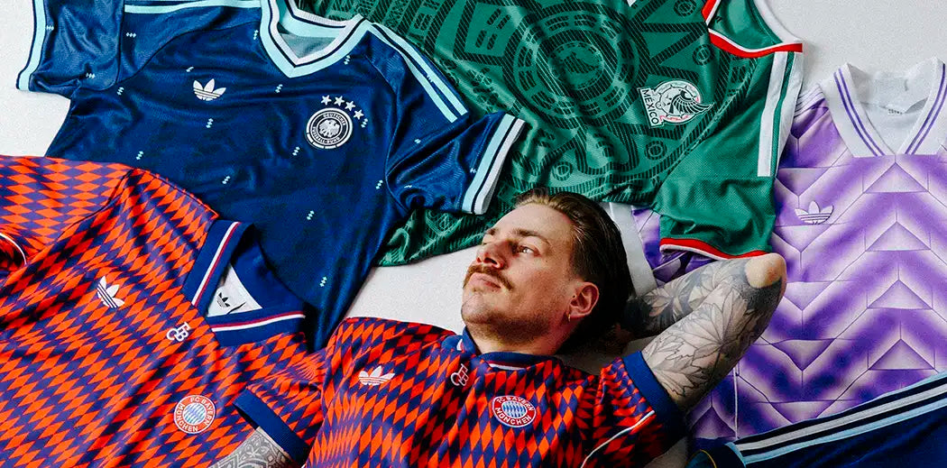 Man with tattooed arm lies on floor wearing a geometric-patterned soccer jersey, surrounded by vintage jerseys in blue, green, and purple tones.