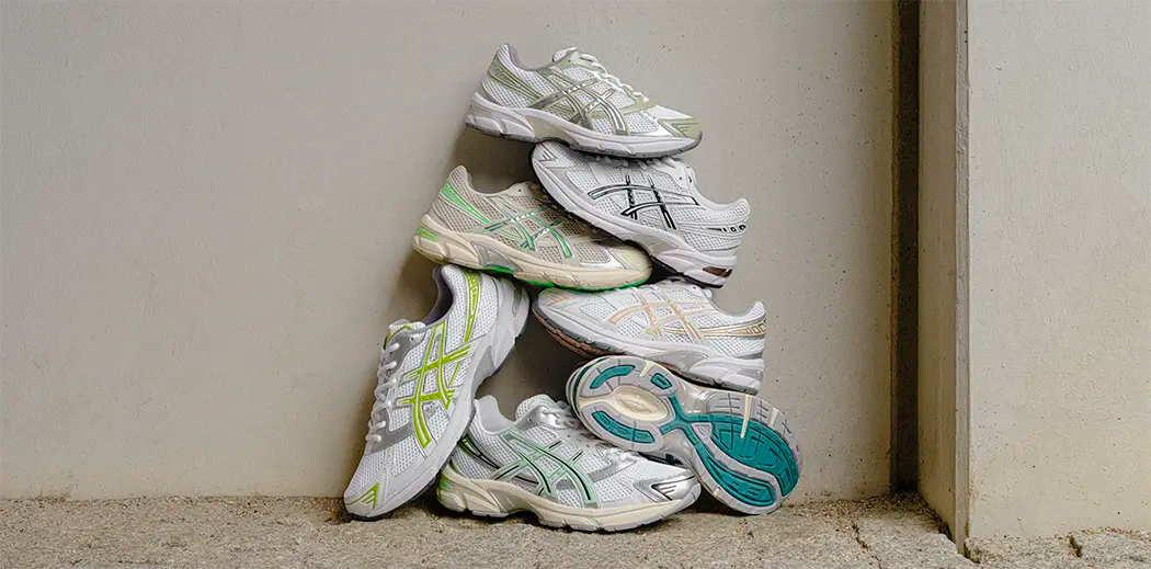 A stack of five white athletic Asics Gel-1130 sneakers with colorful accents, arranged in a pyramid shape against a beige wall. The scene conveys an organized, casual tone.