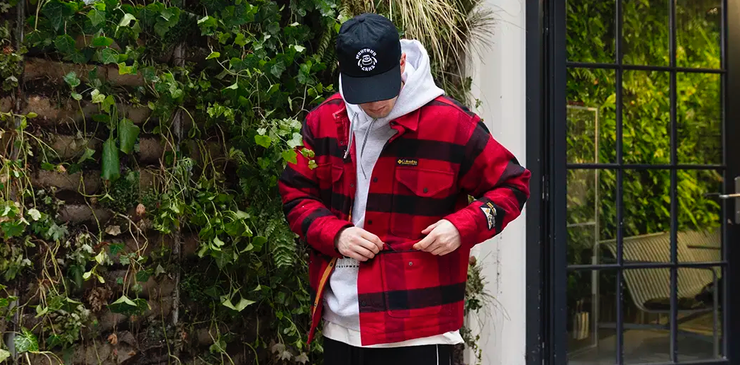A person wearing a red and black plaid jacket over a gray hoodie and a black cap stands near a lush green wall. The mood is casual and urban.