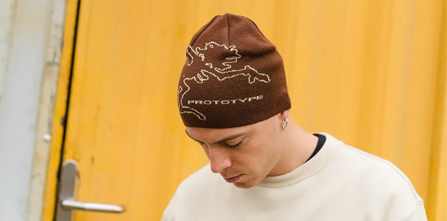 A person with a neutral expression wears a brown beanie with a lion graphic and "PROTOTYPE" text on it. Background is a bright yellow door.