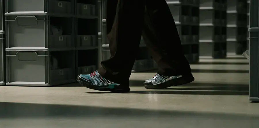 A person walks between rows of shelves filled with grey storage bins wearing a pair of adidas Adistar Control 5 Y2K sneakers. Their colorful sneakers stand out against the neutral, industrial setting.