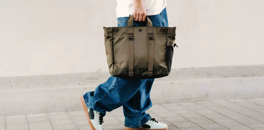 Person in casual attire walks on a sidewalk, carrying an olive green tote bag from Japanese brand Porter Yoshida & Co.. They wear a white shirt, baggy blue jeans, and white sneakers.