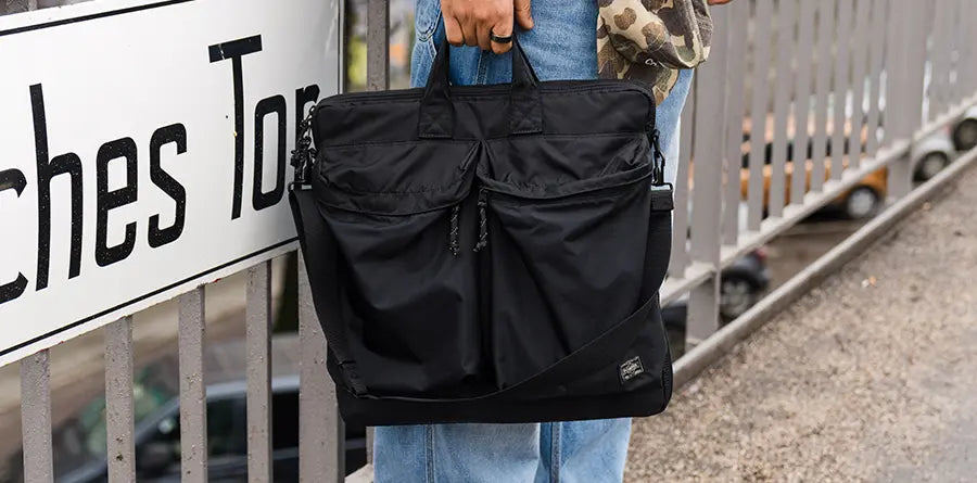A person in jeans holds a black Porter Yoshida & Co. bag with front pockets by a railing. The image has an urban feel, highlighted by a sign partially reading "ches To."
