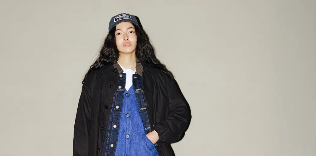 Young person stands confidently against a beige wall, wearing a black coat, blue denim jacket, and jeans. They hold a black bag and wear a cap, exuding a casual, modern vibe. Fallen leaves are scattered on the ground.