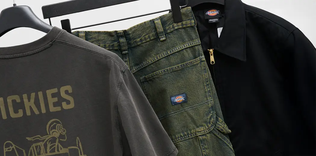 Three pieces of Dickies clothing hang on a rack: a dark graphic tee with "High Flying Workwear," green work pants, and a black jacket, conveying a rugged, durable style.