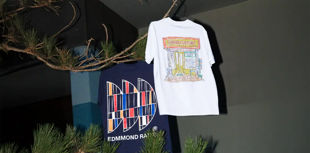 Two T-shirts from Spanish streetwear brand Edmmond Studios hang on a tree branch. The white shirt features a colorful shop illustration, and the navy shirt shows a geometric logo design. The scene feels artistic and casual.