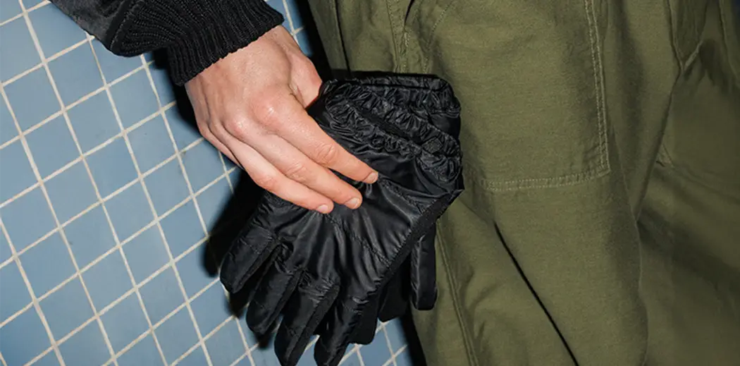 A hand places black gloves into the pocket of olive green pants. The background features a wall of small blue tiles, creating a casual, urban feel.