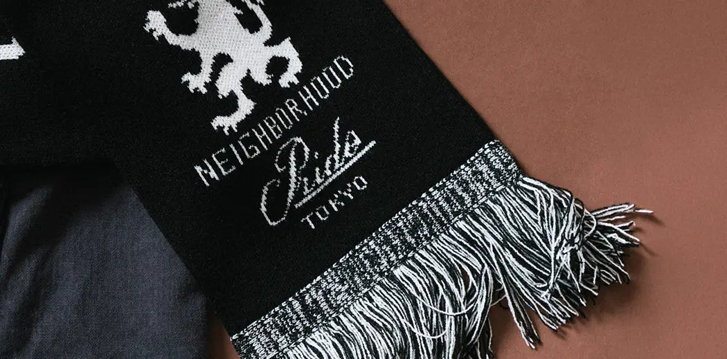 Black scarf with white text and a lion graphic, featuring "NEIGHBORHOOD," "Tokyo," and long fringes. Resting atop a dark fabric on a brown background.