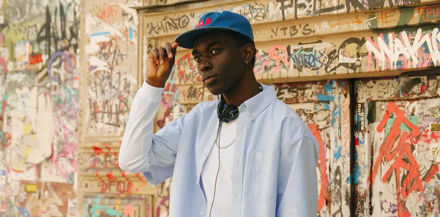 Young man in a blue cap and shirt adjusts his hat while standing against a graffiti-covered wall, exuding a casual and urban vibe.