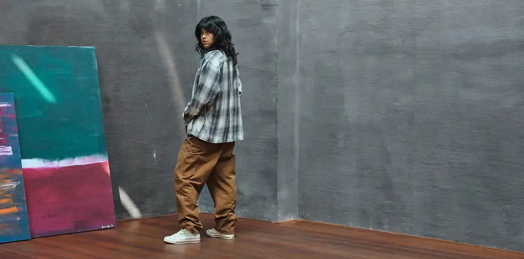 A person with long hair stands on a wooden floor, wearing a plaid shirt and baggy brown pants. They look back towards the camera. A large, colorful painting leans against a gray, textured wall. The scene feels artistic and introspective.
