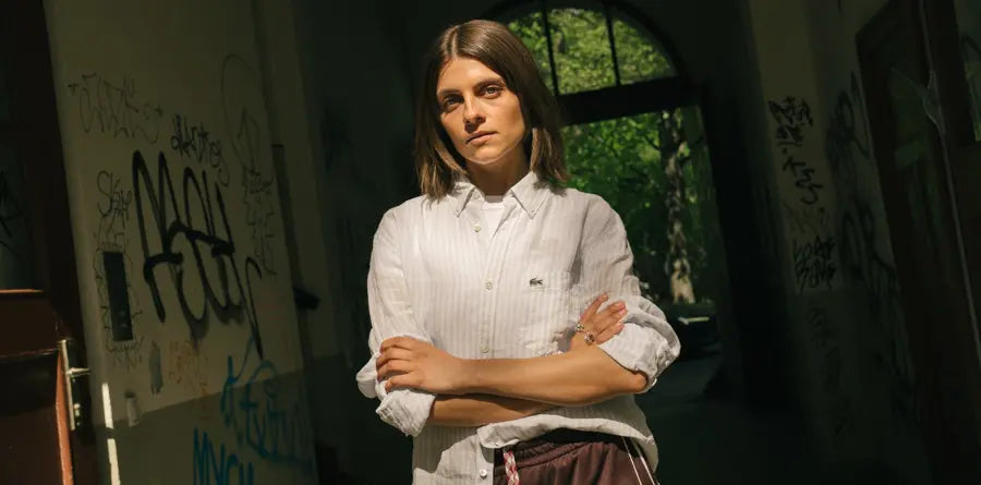 Woman with brown hair in a white shirt stands confidently with arms crossed in a graffiti-covered alley, sunlight and greenery visible in the background.
