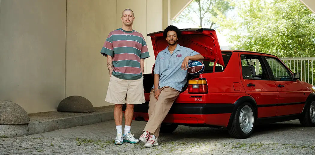 Two men stand by an open car trunk. One wears a striped shirt and beige shorts, the other a blue shirt and wears pants holding a basketball. Casual, relaxed scene.