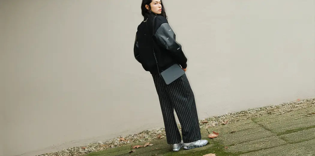A person stands outdoors against a plain wall, wearing a black jacket and pinstripe pants. They hold a small black bag, exuding a confident, relaxed demeanor.