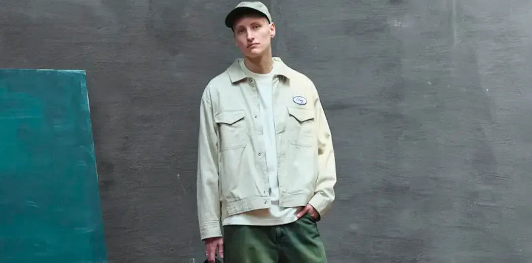 A person wearing a beige jacket, white shirt, and green pants leans against a gray wall. They wear a matching cap, exuding a relaxed, casual vibe.