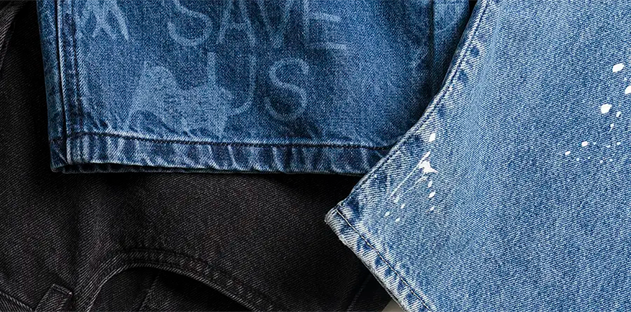 Close-up of overlapping jeans denim fabrics: black, a faded blue with "SAVE US" text, and a light blue with white paint splatters, conveying a distressed, artistic vibe.