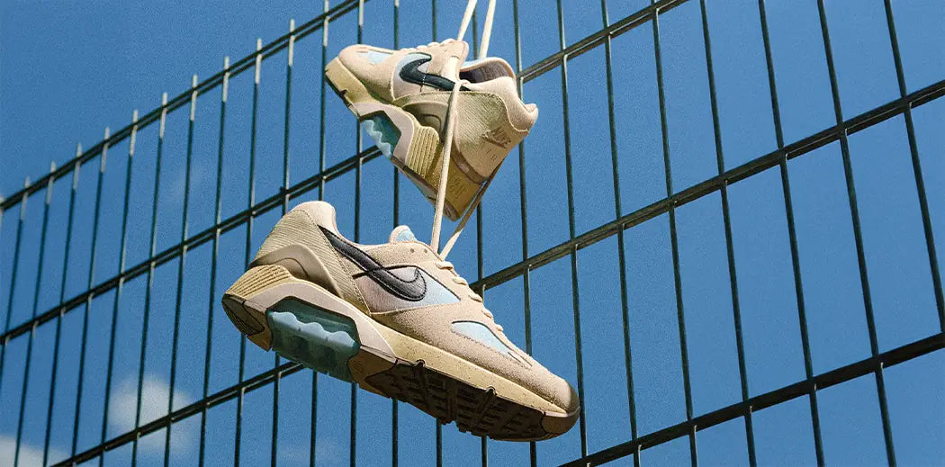 Nike Air Max 180 Sneakers hanging by laces on a green metal fence, with a clear blue sky background. The scene conveys a casual, carefree vibe.