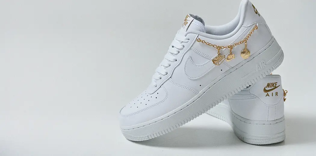 White Nike Air Force 1 '07 LX sneakers adorned with gold charms and text, displayed against a gray background, priced at 129 euros.