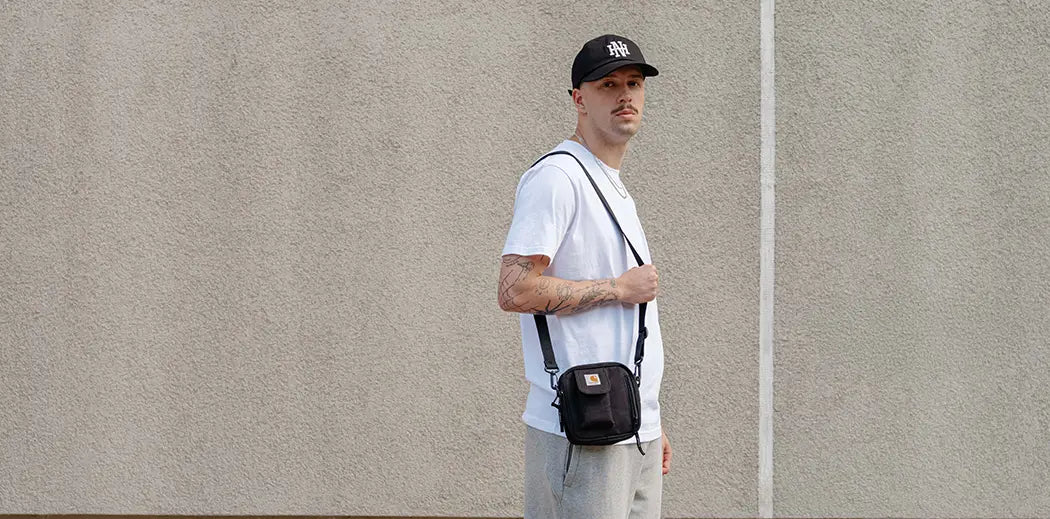 A man stands against a plain beige wall, wearing a black cap, white T-shirt, gray sweatpants, and sneakers, with a small black crossbody bag. The mood is casual.