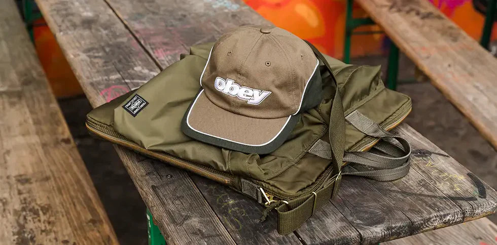 A khaki cap with "obey" text rests on a matching olive green bag on a weathered wooden bench, exuding an urban, laid-back vibe.
