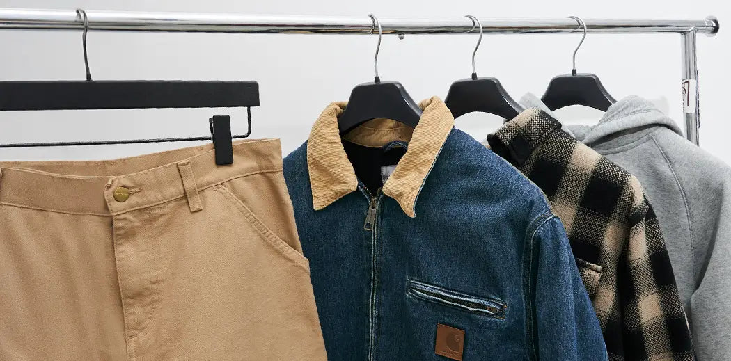Clothes hanging on a rack: beige pants, a denim jacket with tan collar, a checkered flannel shirt, and a gray hoodie. Casual, stylish tone.