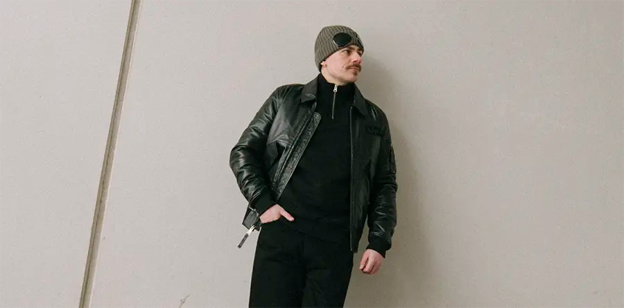 Man in a leather jacket and knit beanie leans against a light wall, hands in pockets, looking to the side. The tone is relaxed and stylish.