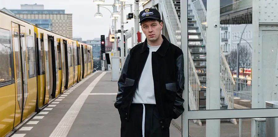 Young man in a black jacket and cap stands on an urban train platform with a yellow train in the background. The mood is relaxed and contemplative.