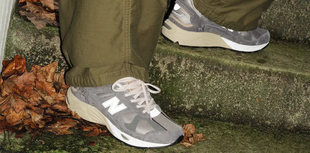 A pair of gray New Balance 878 sneakers with white laces on mossy concrete steps surrounded by dried leaves. The wearer is in olive green pants. Autumn tones.