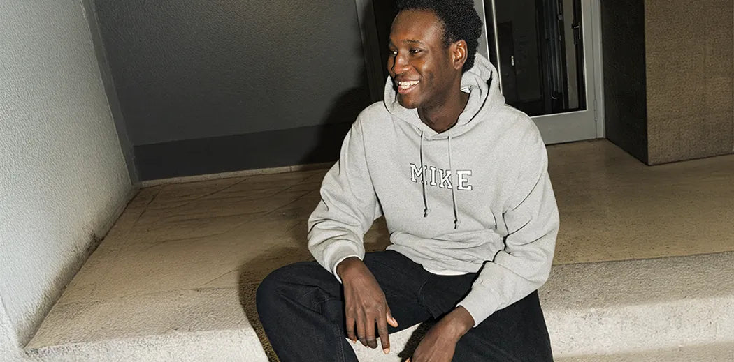 A person sits on steps, smiling and wearing a gray hoodie with "MIKE" text, paired with black pants. The setting is urban, with a relaxed atmosphere.