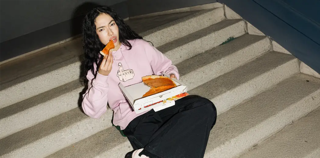Female person in a pink hoodie eating pizza, sitting on stairs. The hoodie has an illustrated design, and they appear relaxed and casual.