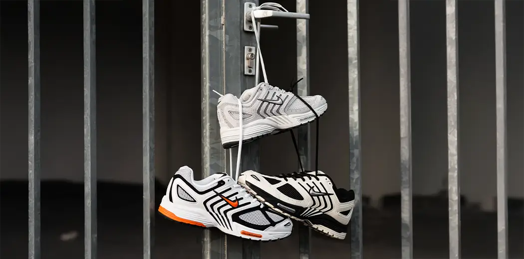 Three Nike sneakers hang by their laces on a gray metal gate. The shoes are white, black, and orange, creating a casual, artistic display.