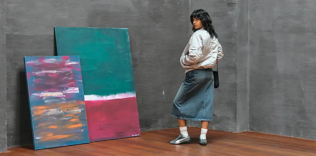 A person in a silver jacket and denim skirt observes two large abstract paintings with vibrant colors, leaning against a gray wall. The setting feels contemplative.