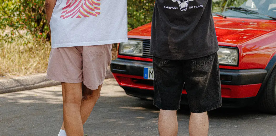 Two men stand with their backs to the camera, one in a white "ARTE" shirt and pink shorts, the other in a black graphic tee. A red car and trees are in the background.