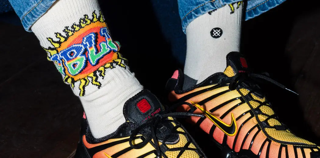 Denim-clad person sits on a dark surface, wearing vibrant orange and yellow Nike sneakers with unique soles and colorful socks from STANCE.