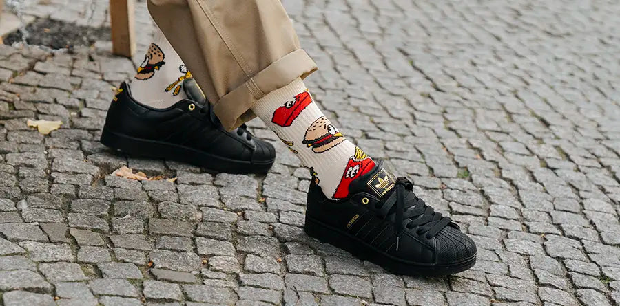 Person wearing beige pants, white STANCE socks with cartoon burger and fries patterns, and black sneakers standing on cobblestone pavement