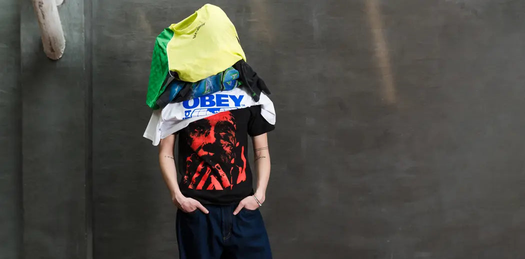 Person stands against a gray wall with multiple men T-shirts stacked over their head, obscuring their face, hands in pockets. The scene is quirky and playful.