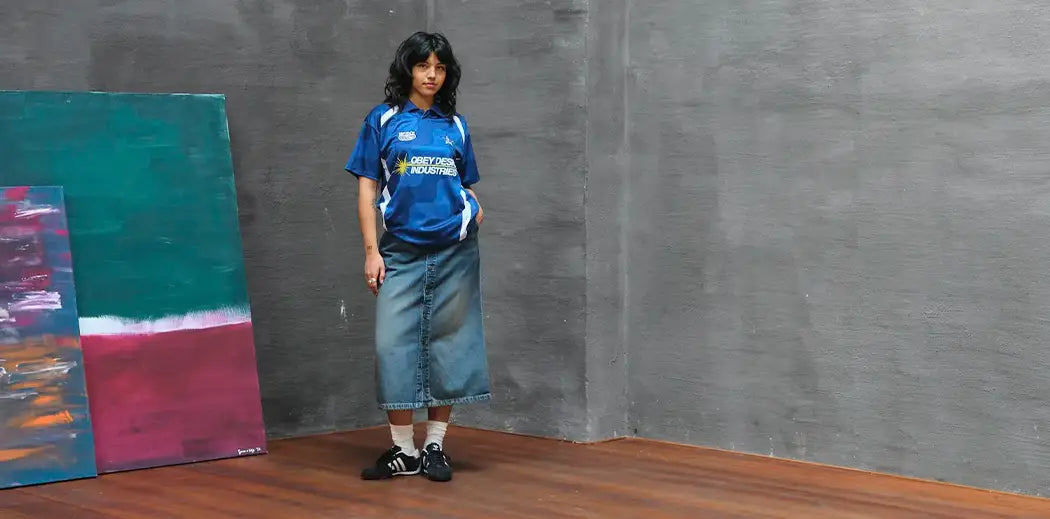 A person in a blue sports jersey and denim skirt stands indoors against a gray wall beside colorful abstract paintings. The scene is calm and artistic.