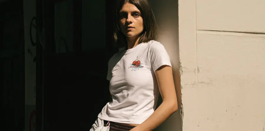 A woman in a white t-shirt with a cherry design stands against a sunlit wall, casting shadows. She gazes confidently, creating a moody, relaxed atmosphere.