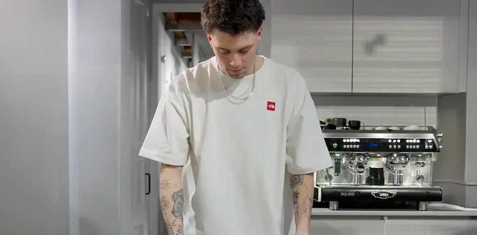 A person wearing a white T-shirt stands in a modern kitchen, looking down. Tattoos are visible on their arms. An espresso machine is in the background.