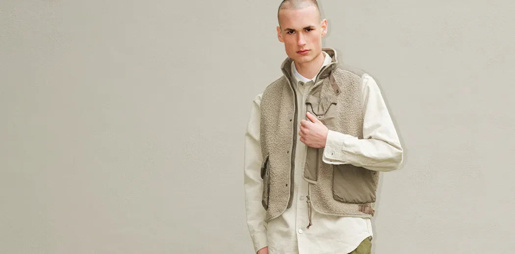 A person stands against a beige wall, wearing a light shirt, beige vest, green pants, and sneakers. The ground is covered with moss and fallen leaves, conveying an autumnal tone.