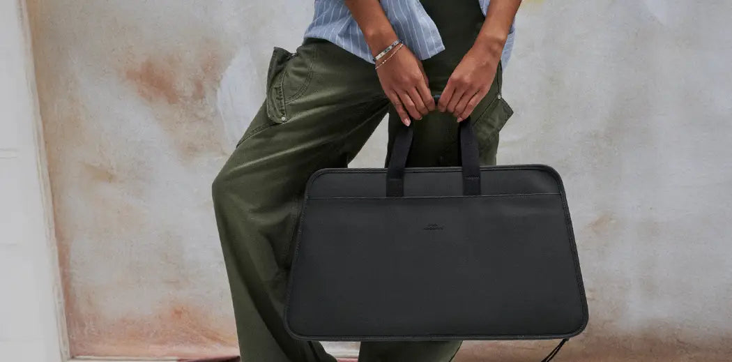 A person in a striped blue shirt and olive cargo pants holds a large black bag. Their stance is relaxed against a subtle, artistic background.