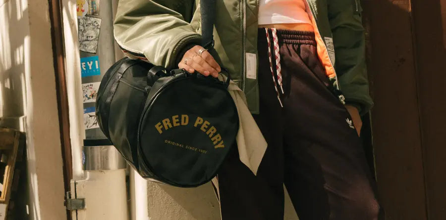 A person holds a black Fred Perry duffle bag against their side. They wear a green jacket and black pants. The setting feels urban and casual.