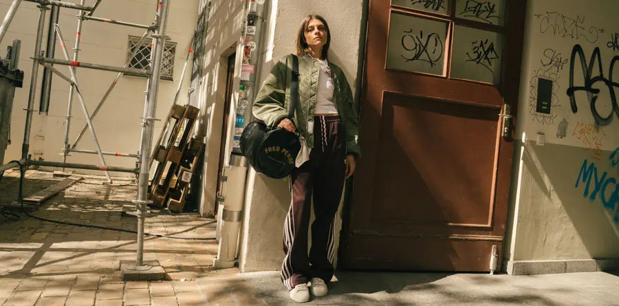 A woman in casual streetwear stands in a sunlit urban alley. She wears a green bomber jacket, track pants, and sneakers, holding a duffel bag. The wall behind her is covered with graffiti.