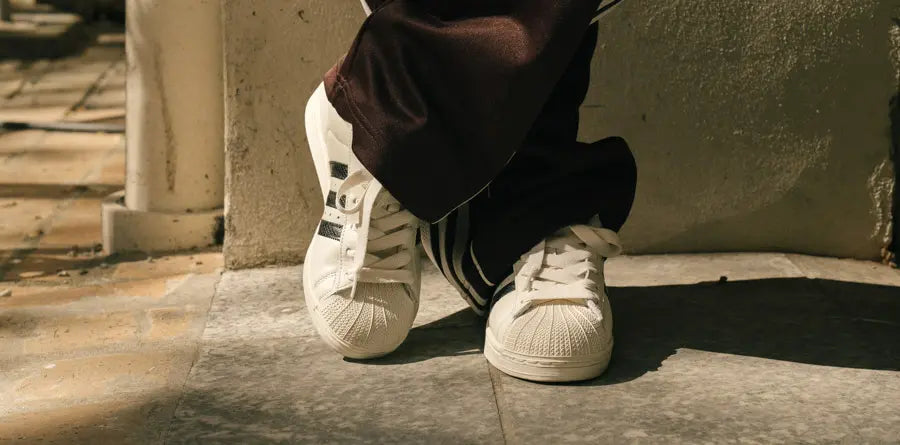 Sneakers with black stripes on a person standing with crossed legs. The scene conveys a relaxed mood with sunlight casting soft shadows on a tiled floor.