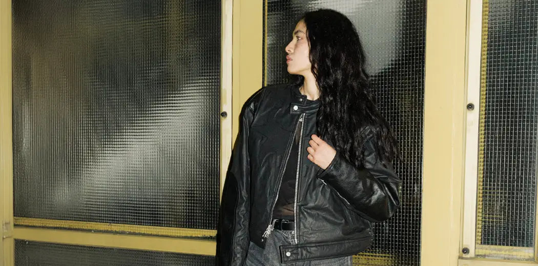 A person in a black leather jacket and wide-legged jeans leans casually against a yellow, textured glass door, exuding a cool and relaxed vibe.