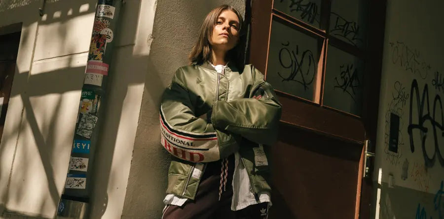 A woman stands confidently, arms crossed, wearing a green bomber jacket. She's against a graffiti-covered wall with a serious expression, exuding urban style.
