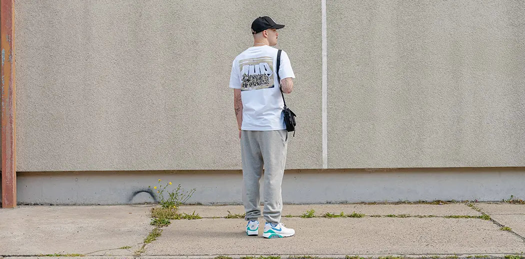 A person stands facing a beige concrete wall, wearing a white T-shirt from the graffiti crew 1UP OneUP, gray pants, a black cap, and a crossbody bag. The scene is calm and minimalist.