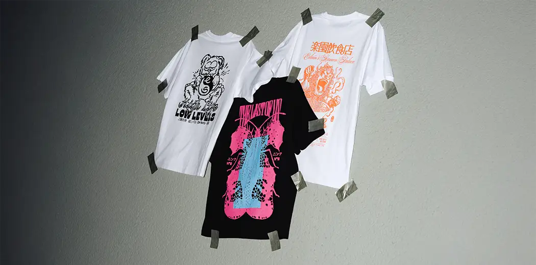 Three graphic T-shirts from Japanese brand Edwin taped to a textured wall. Designs include a cartoon bear, vibrant abstract shapes, and Asian characters. Creative and edgy vibe.