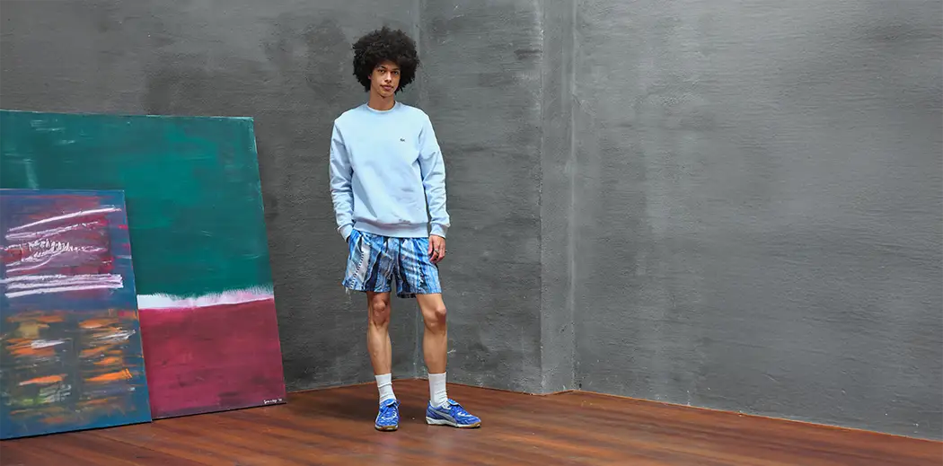 A person stands confidently in an art studio, wearing a light blue sweater from Lacoste and plaid shorts. Colorful abstract paintings lean against a gray wall.