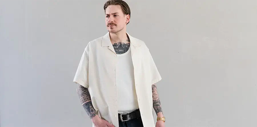 A man with tattoos and a mustache stands confidently. He's wearing a white open shirt from Lacoste over a tank top, black pants, and a watch, exuding a relaxed vibe.